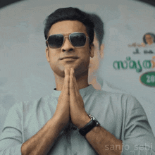 a man wearing sunglasses holds his hands together in front of a sign that says sanjo_sebi
