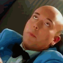 a bald man wearing a blue suit and bow tie looks up