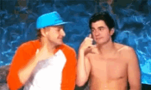 a shirtless man in a blue hat talks to another shirtless man in an orange jacket