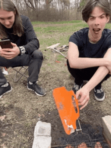a man squatting next to another man who is holding an orange spatula that says ' s' on it