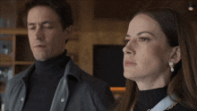 a woman wearing pearl earrings stands next to a man wearing a turtleneck
