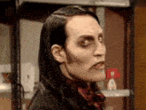 a man with long black hair and makeup is standing in front of a shelf and making a funny face .