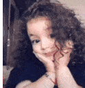 a little girl with curly hair is smiling with her hand on her chin .