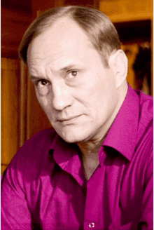 a man in a pink shirt looks at the camera with a serious look on his face