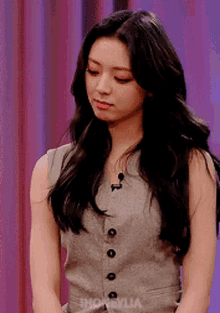 a woman with long black hair is wearing a grey vest and a purple curtain behind her .