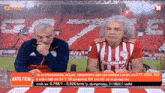 two men are sitting in front of a soccer field and one has a shirt that says stoiximan on it