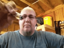 a bald man with a beard is wearing glasses and a blue shirt in a garage .