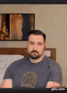 a man with a beard is sitting in front of a bed wearing headphones and a t-shirt .