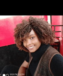 a woman wearing a black turtleneck and a brown vest is smiling for the camera on a redmi note 6 pro