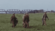 a group of soldiers walking through a grassy field with a bridge in the background