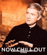 a man in a black shirt is sitting in front of a fire with his hands out .