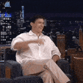 a man in a white shirt is sitting in a chair with his legs crossed and smiling .