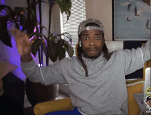 a man with dreadlocks wearing a hat and a grey sweatshirt with the letter t on the front