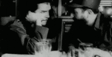 a black and white photo of two men sitting at a table talking .