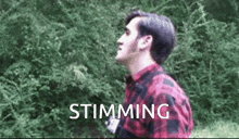 a man in a red and black plaid shirt is standing in front of trees with the word stimming written on the bottom .