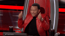a man in a red jacket is sitting in a chair with a nbc logo in the background