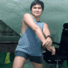 a man in a blue tank top and shorts is dancing in a room with a chair .