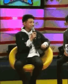 a man in a black jacket is sitting in a yellow chair holding a microphone