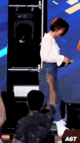 a woman in a white shirt and blue jeans stands in front of a sign that says agt on it