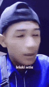a man wearing a hat and a blue shirt says lelaki setia on the bottom of his face .