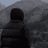 a person in a hooded jacket stands in front of a mountain range