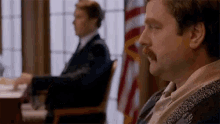 a man with a mustache is sitting in a chair in front of a flag .