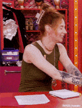 a woman with tattoos on her arms sits at a desk with a clipboard