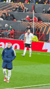 a man in a blue jacket stands on a soccer field in front of an ad for adidas