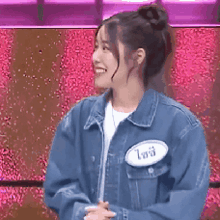 a woman wearing a denim jacket and a name tag is standing in front of a pink background .