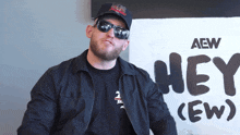 a man wearing sunglasses and a hat stands in front of a sign that says hey ( ew )