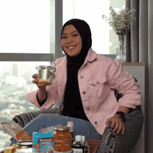 a woman in a pink jacket is sitting in a chair holding a jar and smiling