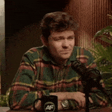 a man in a plaid shirt is sitting at a table with a microphone in front of him .