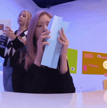a woman covering her face with a piece of paper while another woman looks on