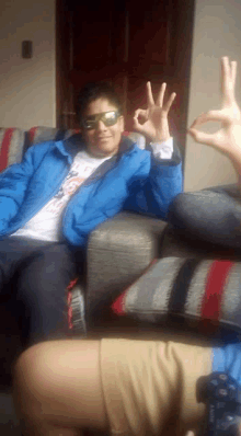 a man wearing sunglasses and a blue jacket sitting on a couch giving the ok sign