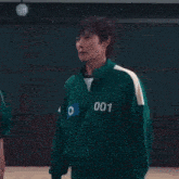 a man in a green jacket with the number 001 on it is standing in front of a group of people in a gym .