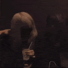 a woman with blonde hair is sitting in a dark room holding a cup with a straw