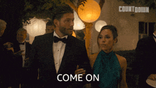 a man in a tuxedo and a woman in a green dress are standing next to each other and the woman is saying come on