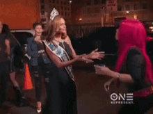two women are sitting next to each other on a couch . one of the women is wearing a pink wig .