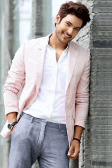 a man wearing a pink jacket and grey pants smiles while leaning against a wall