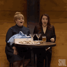 two women sit at a table with martini glasses and oysters and a snl logo in the corner