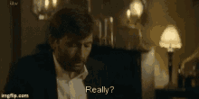 a man in a suit and white shirt is sitting in a dark room with a lamp and says `` really '' .