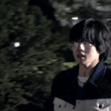 a young man wearing glasses and a blue jacket is standing in a forest .