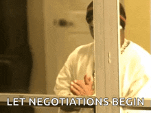 a man is standing in front of a window with his hands folded and the words `` let negotiations begin '' written on the window .