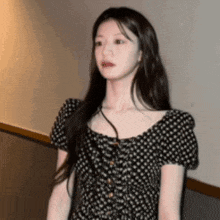 a woman in a black and white polka dot dress is standing next to a wall and looking at the camera .