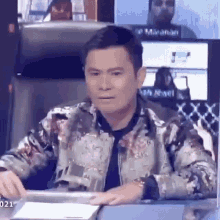 a man in a floral jacket and tie is sitting at a desk in front of a computer .
