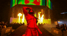 a wrestler in a red outfit is walking down a stage with fire coming out of the ceiling .