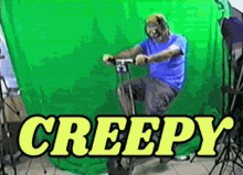 a man in a blue shirt is riding an exercise bike in front of a green screen that says creepy in yellow letters