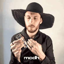 a man wearing a black hat is holding a glass with the word mcdh on the bottom right