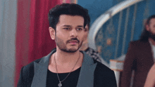 a man with a beard and a necklace is standing in a room looking at the camera .