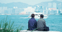 two men sit on a ledge overlooking a body of water with a boat in the distance